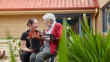 Female home care worker holding a pot plant and kneeling next to female Home Care Package client sitting on a walker